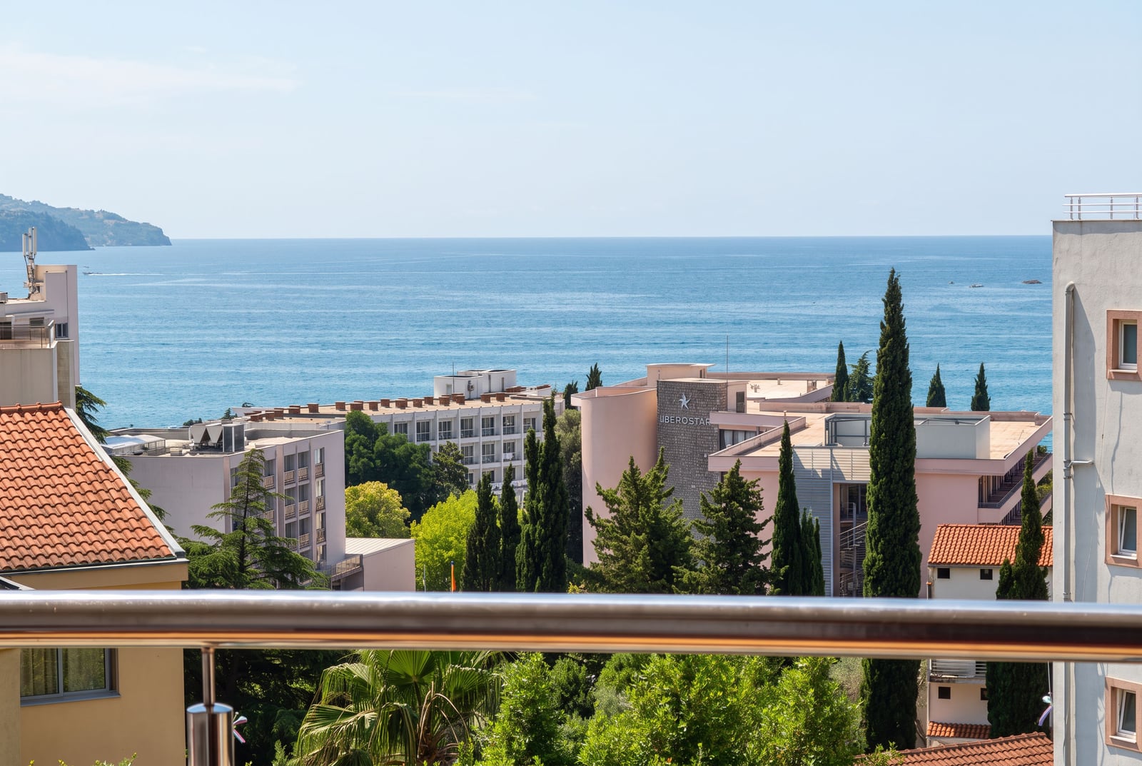 View from a balcony overlooking the sea, buildings, and trees. Metal railing is visible in the foreground.