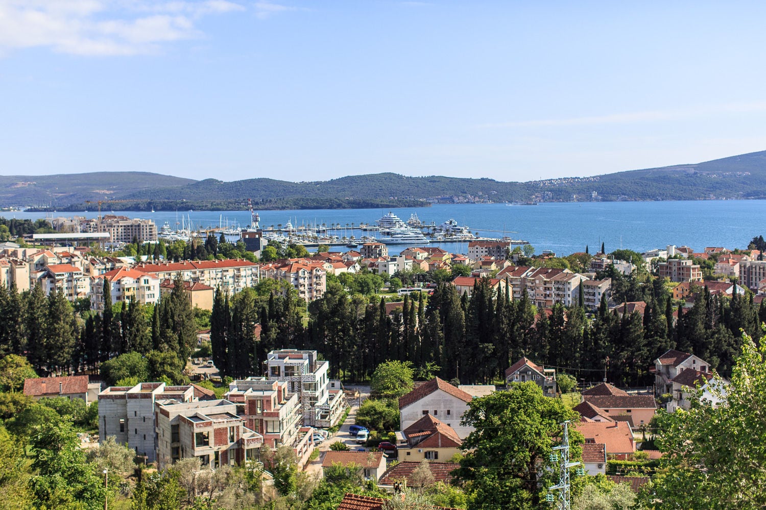Panoramic exterior view of a coastal town with red roofs, a marina filled with boats, and sea.