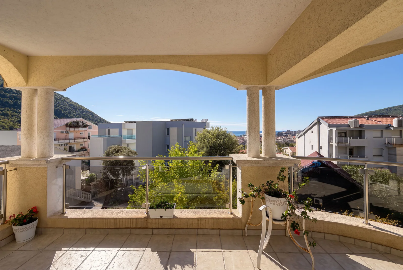 Tiled balcony with glass railing and archway, overlooking city and sea, potted plants visible.