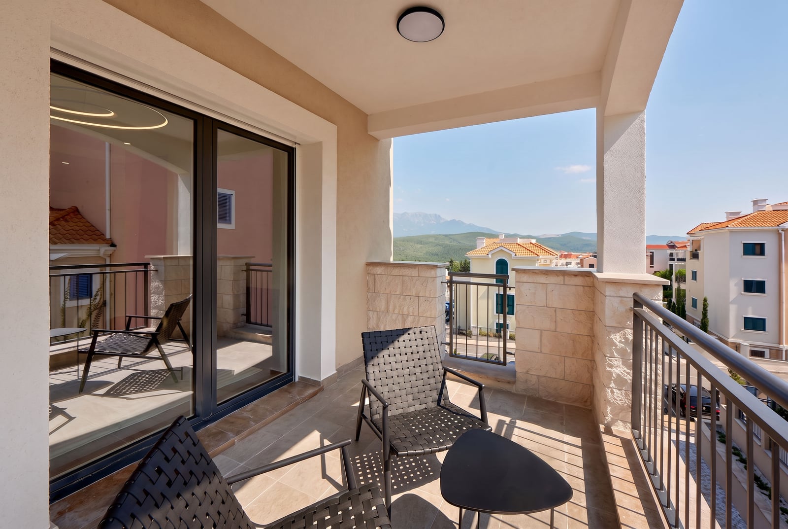 Open balcony with woven chairs and table, overlooking mountains and neighboring houses through a metal railing.