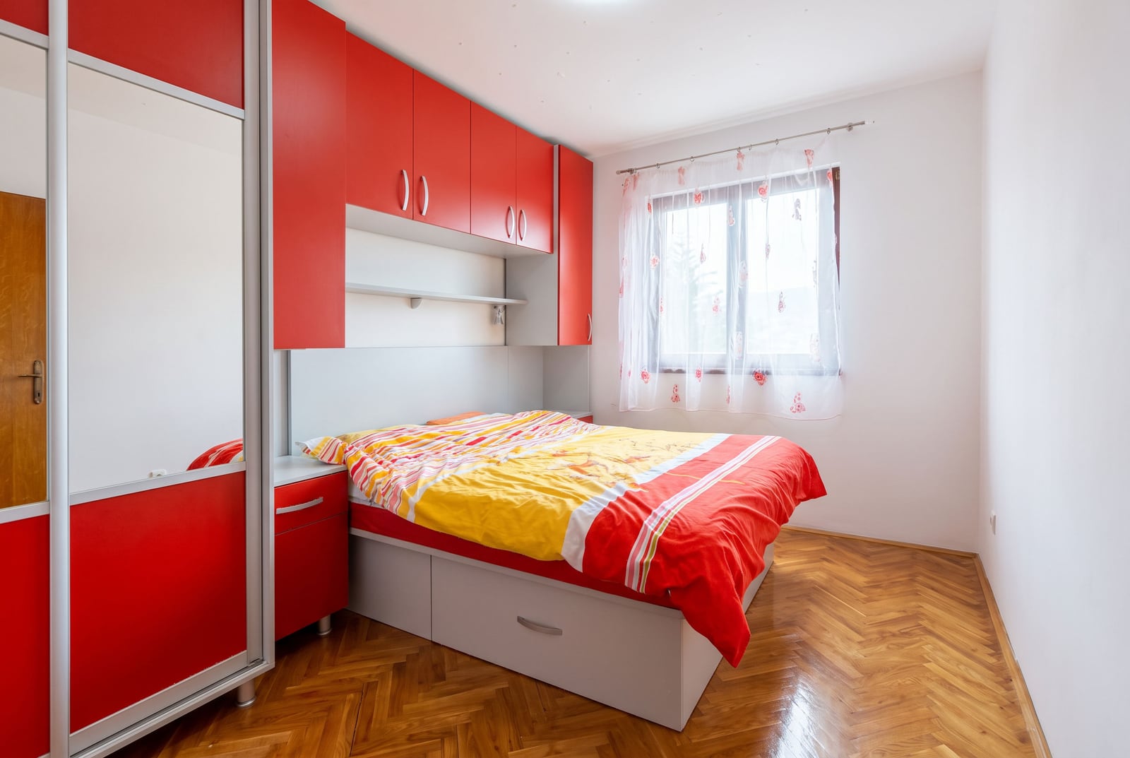 Bedroom featuring red built-in cabinets, a bed with colorful bedding, and a mirrored wardrobe on parquet flooring.