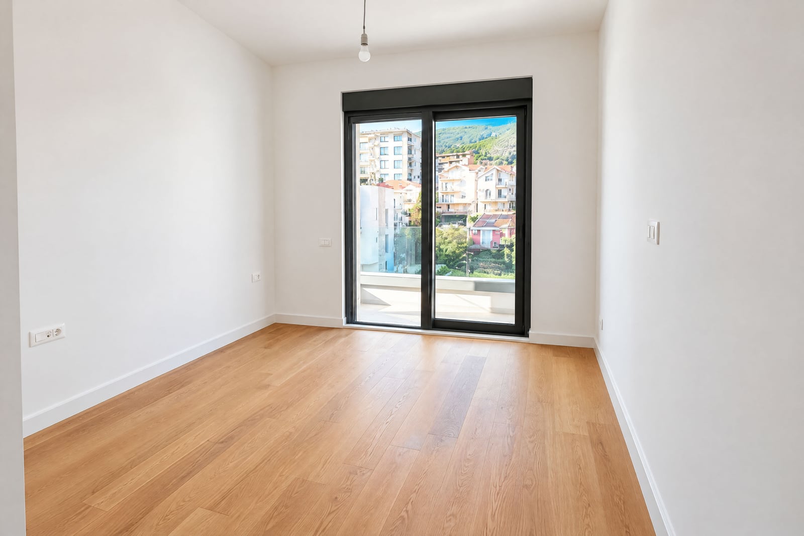 Empty room with wooden flooring and white walls, featuring sliding glass doors leading to a balcony.