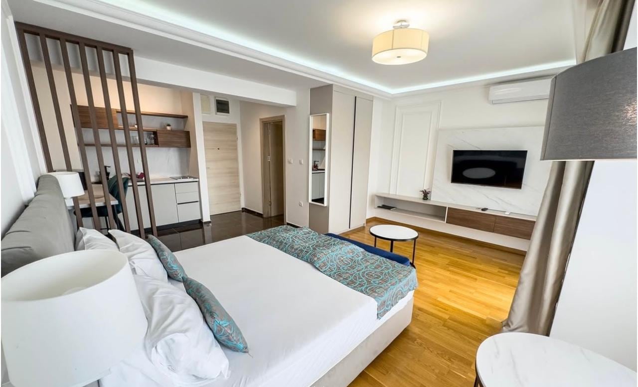 Modern bedroom in an apartment in Bečići, Montenegro: double bed, kitchen area, and wood-paneled TV wall.