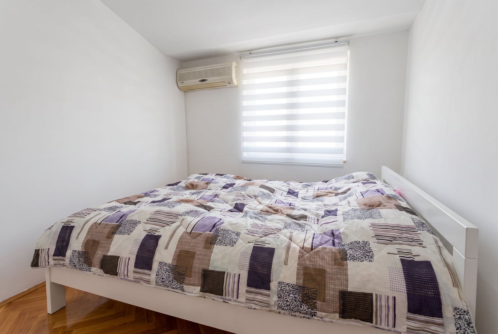 Bedroom featuring a white bed with patchwork bedding, wall-mounted AC unit, window with blinds, and parquet flooring.