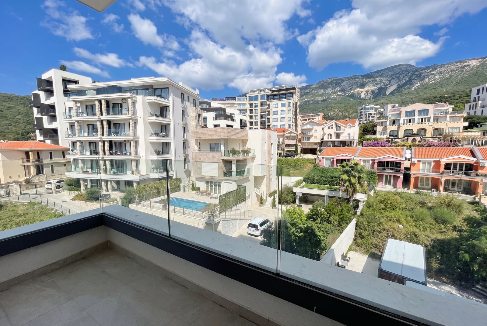 View from a balcony with glass railing overlooking residential buildings, a pool, and mountains under a cloudy sky.