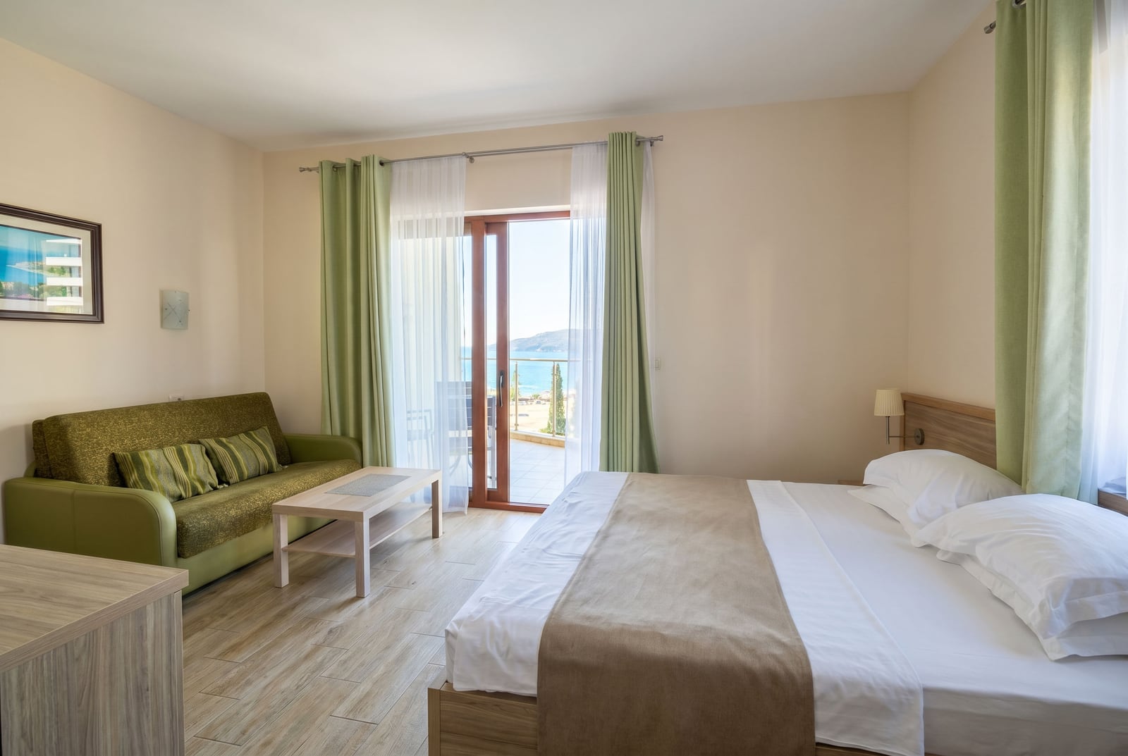 Bedroom in an apartment in Becici, Montenegro: double bed, green sofa, and access to a terrace with a sea view.