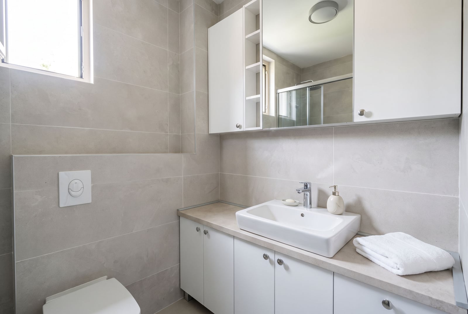 Modern bathroom with grey tiles, white vessel sink on a cabinet, mirrored storage unit, and visible toilet.