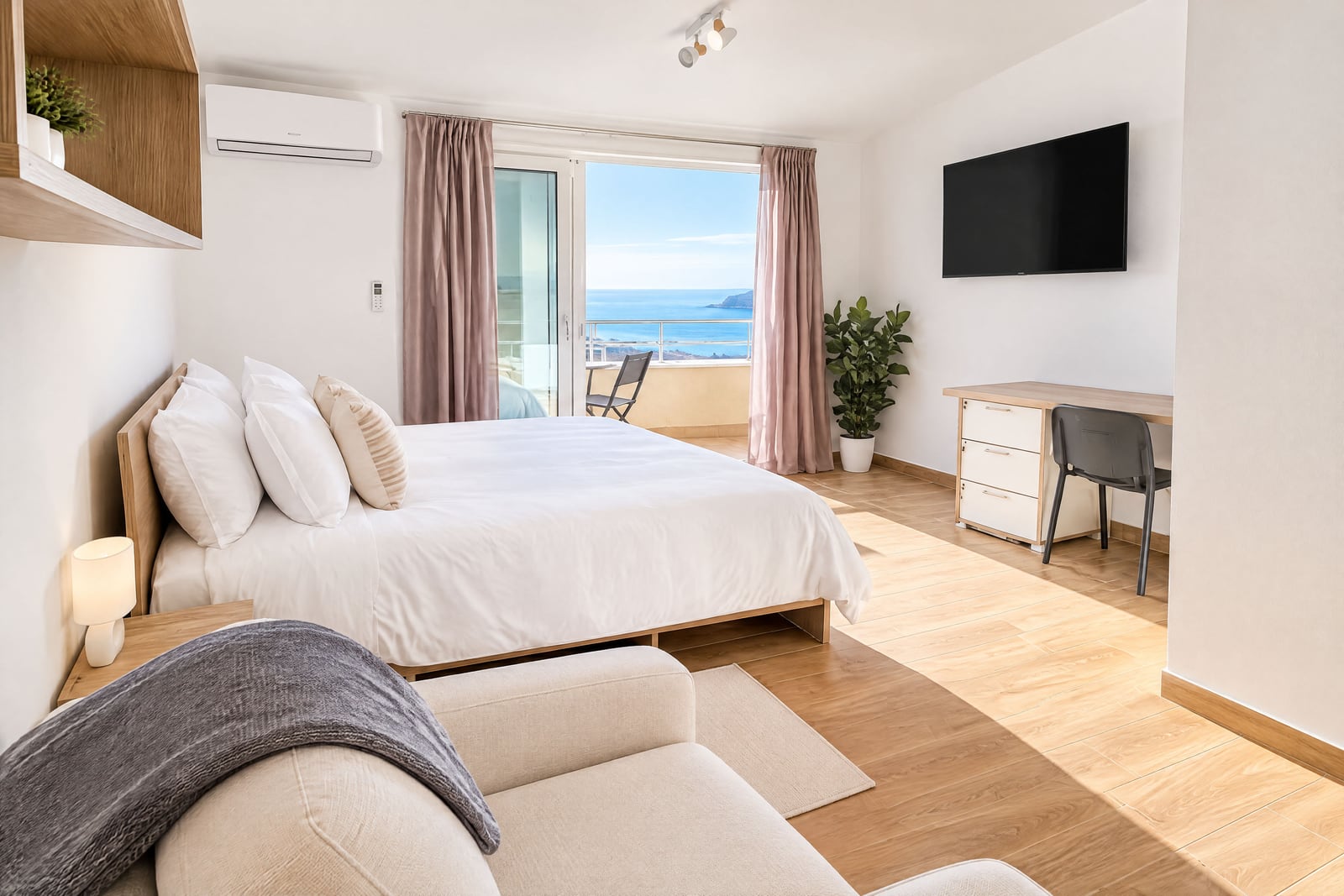 Bright bedroom with a double bed, desk, and balcony door overlooking the ocean.