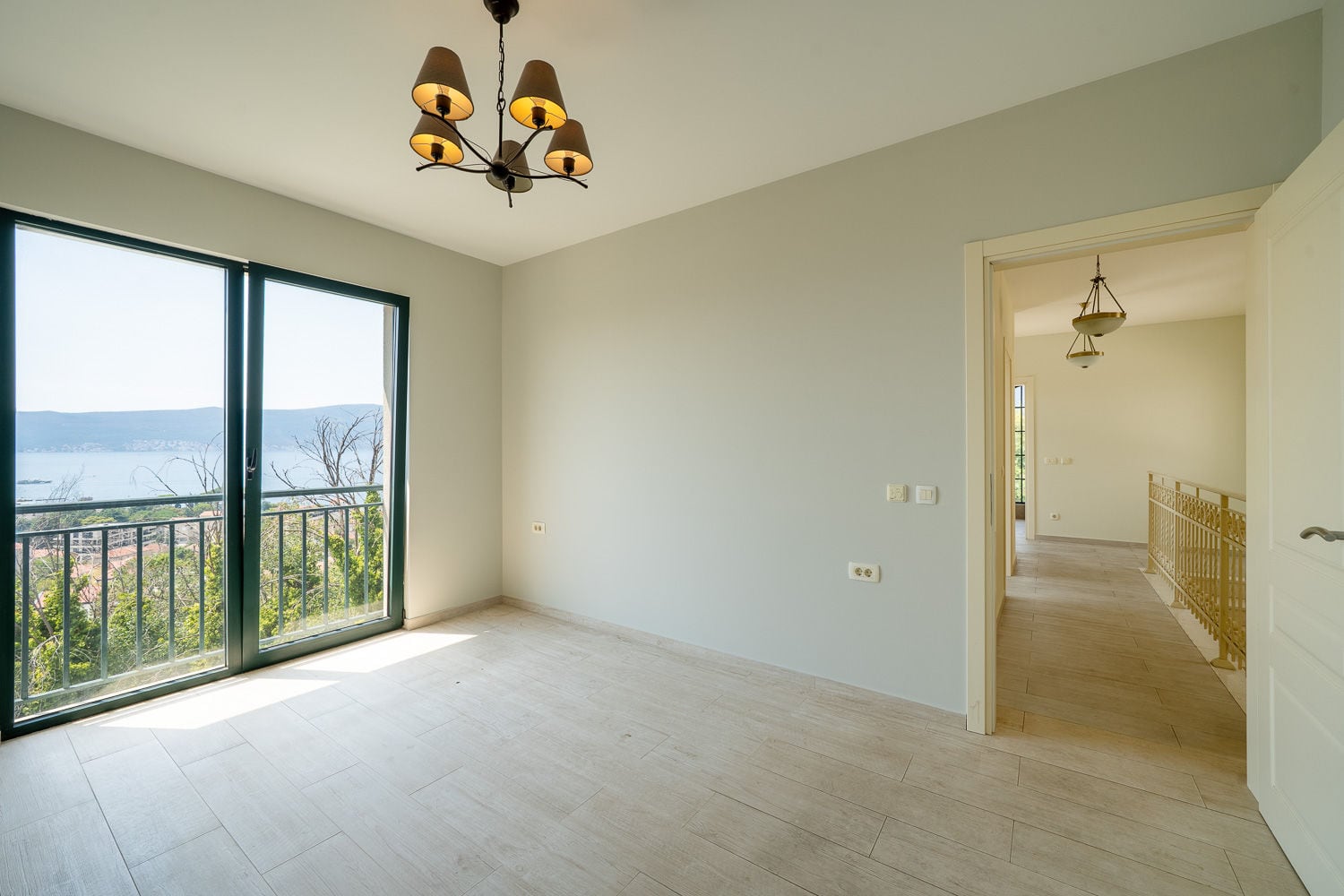 Empty room with light wood flooring, a chandelier, and large glass doors leading to a balcony with a sea view.