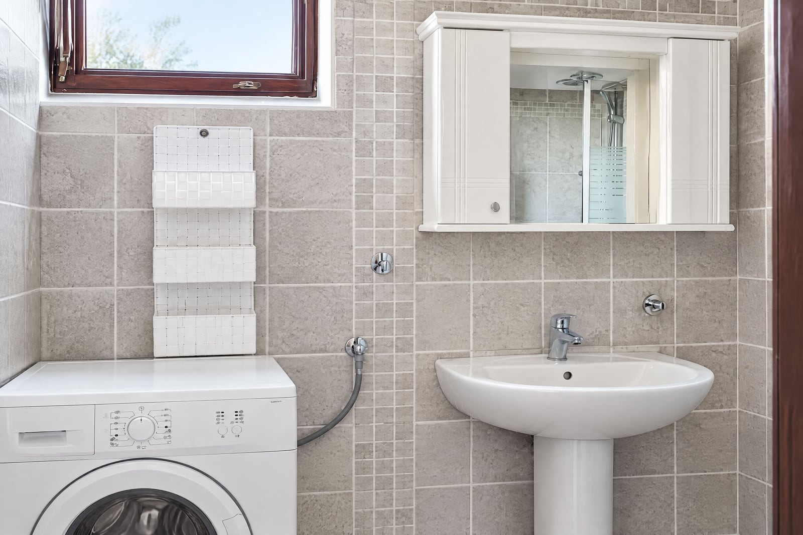 Bathroom with grey tiles, washing machine, pedestal sink, and mirrored storage cabinet.