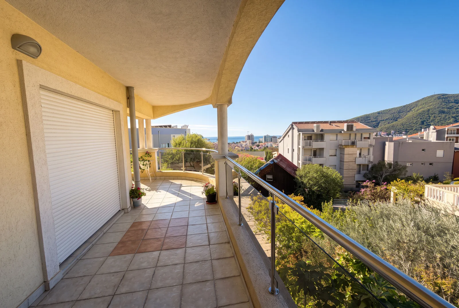 Tiled terrace with metal railing overlooking the sea and mountains. Features a white roll-up door and potted plants.