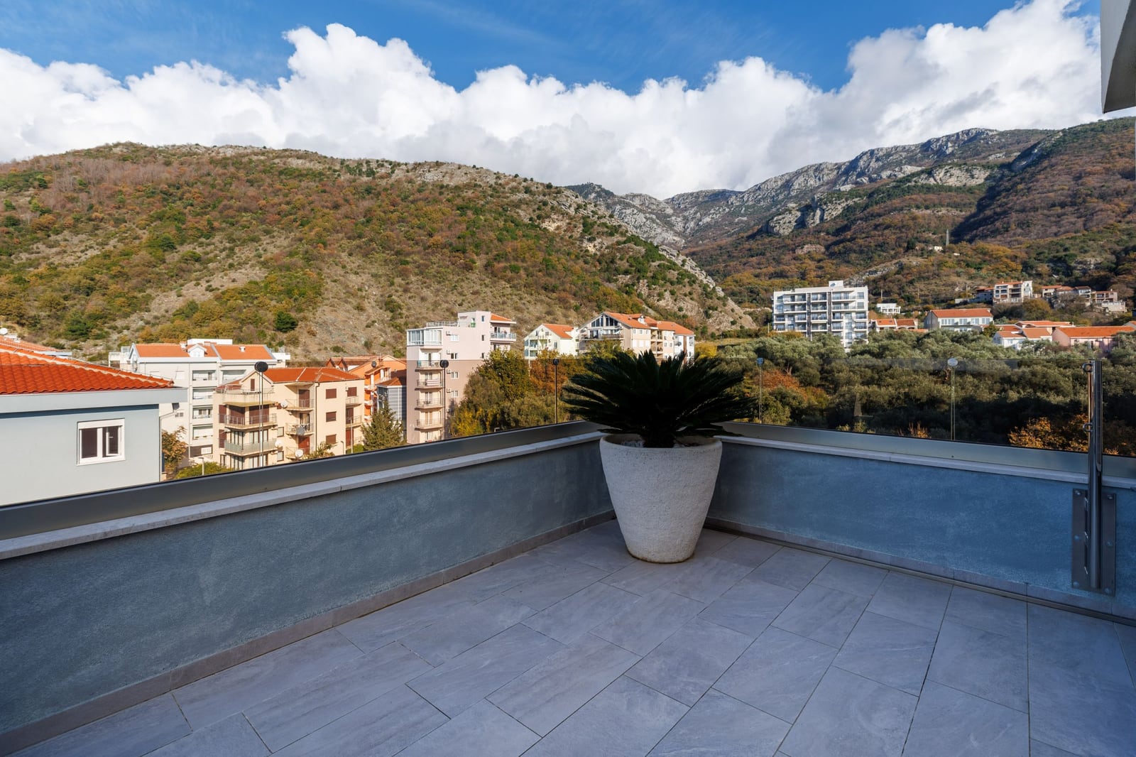 Spacious penthouse terrace in Becici, Montenegro, with panoramic views of the mountains and the city, featuring floor tiles and a potted palm tree.