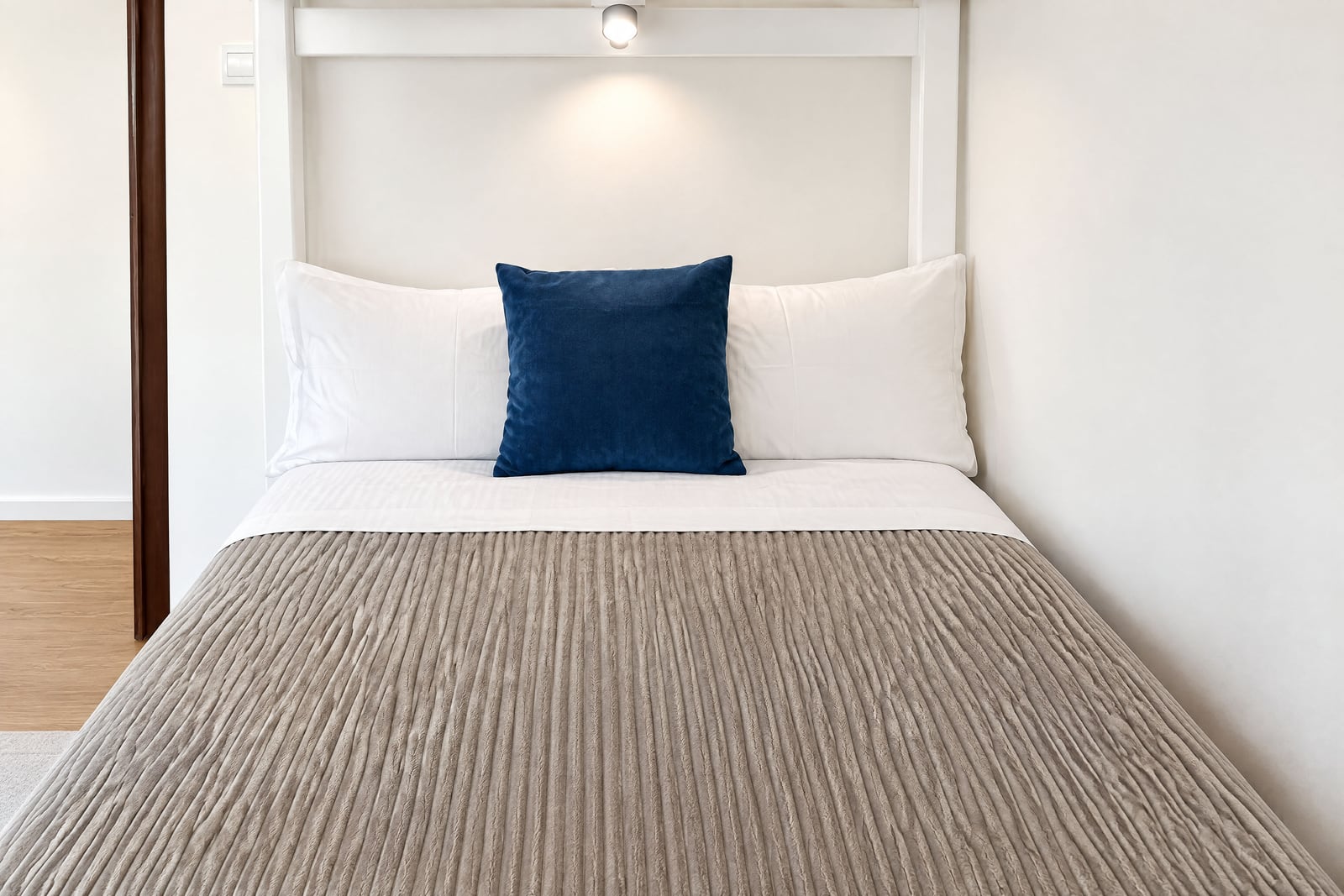 Bed with beige coverlet, white and blue pillows at the headboard under a light fixture.