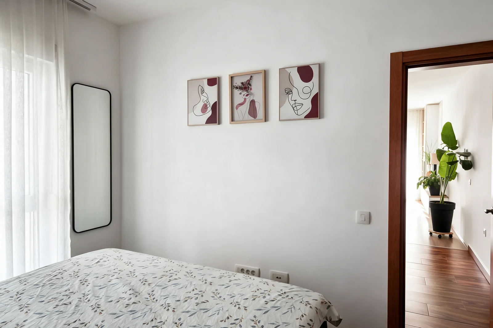 Bedroom with a floral bedspread, tall mirror, and three abstract art prints on a white wall.