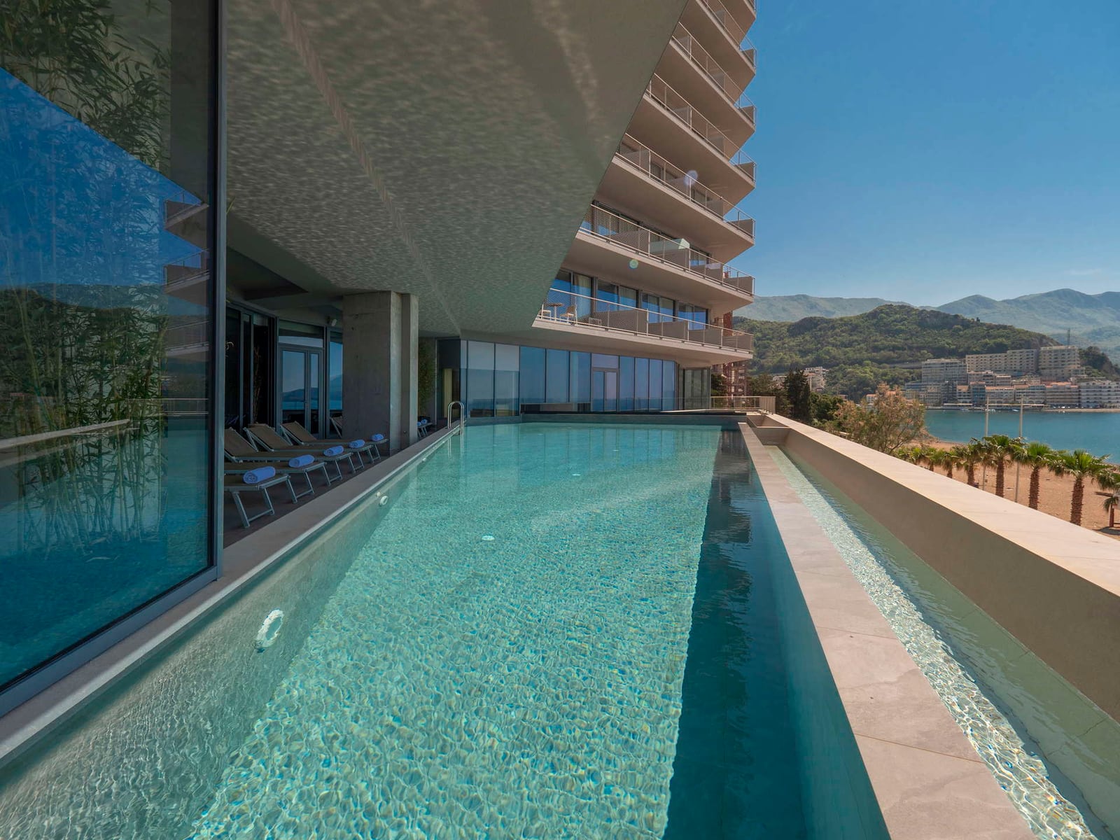 Long rooftop infinity pool overlooking the sea and mountains, with lounge chairs and modern architecture.