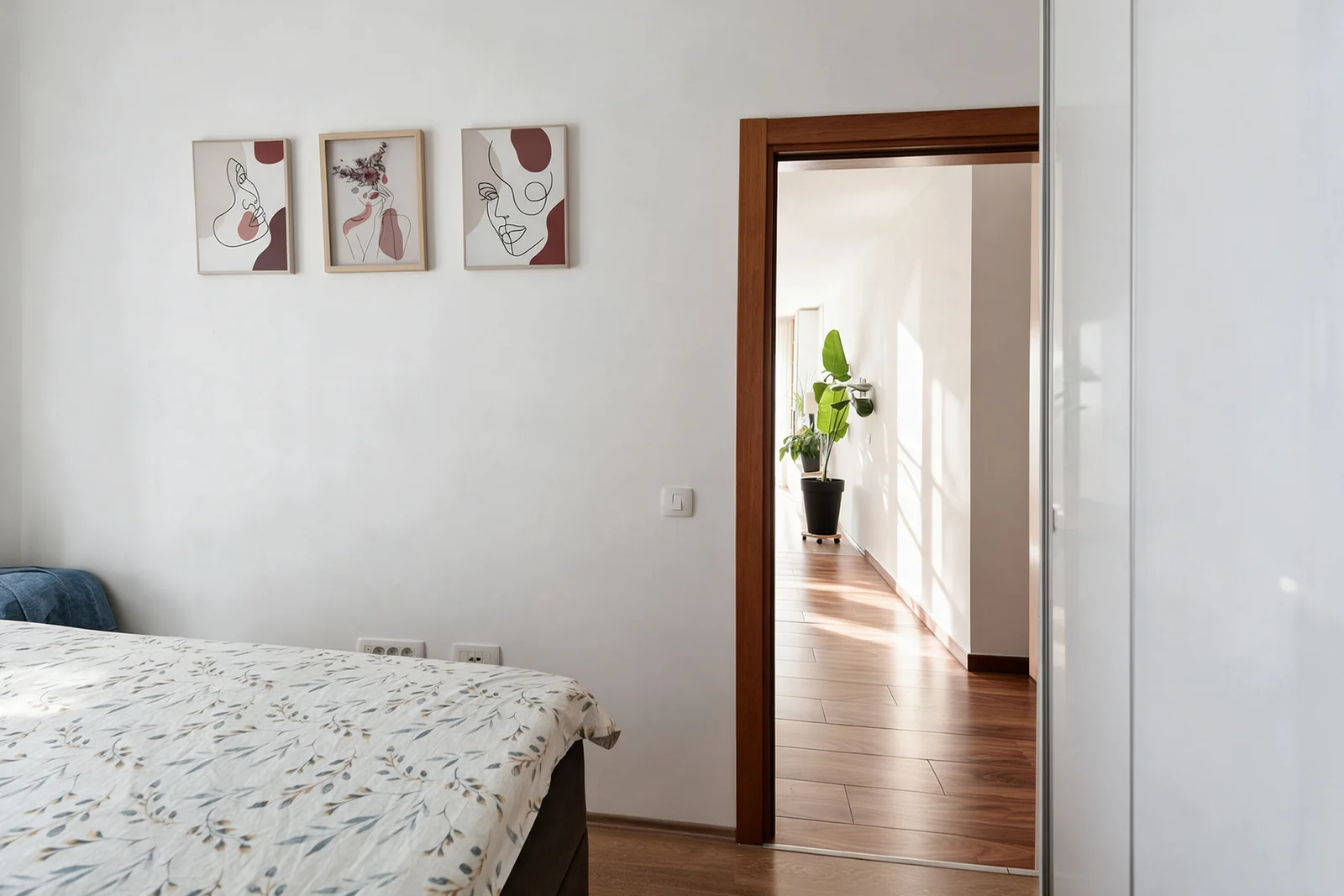 Bed with floral coverlet, three abstract art prints on wall, and doorway leading to a sunlit hallway with a plant.