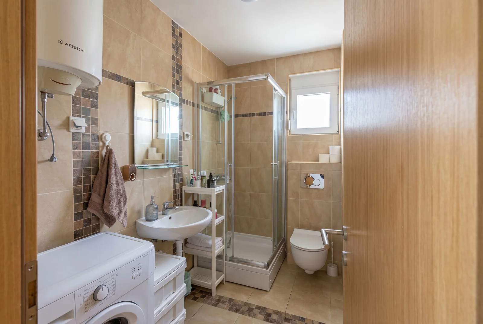 Bathroom featuring a shower stall, sink, wall-mounted toilet, and washing machine on beige tiles.
