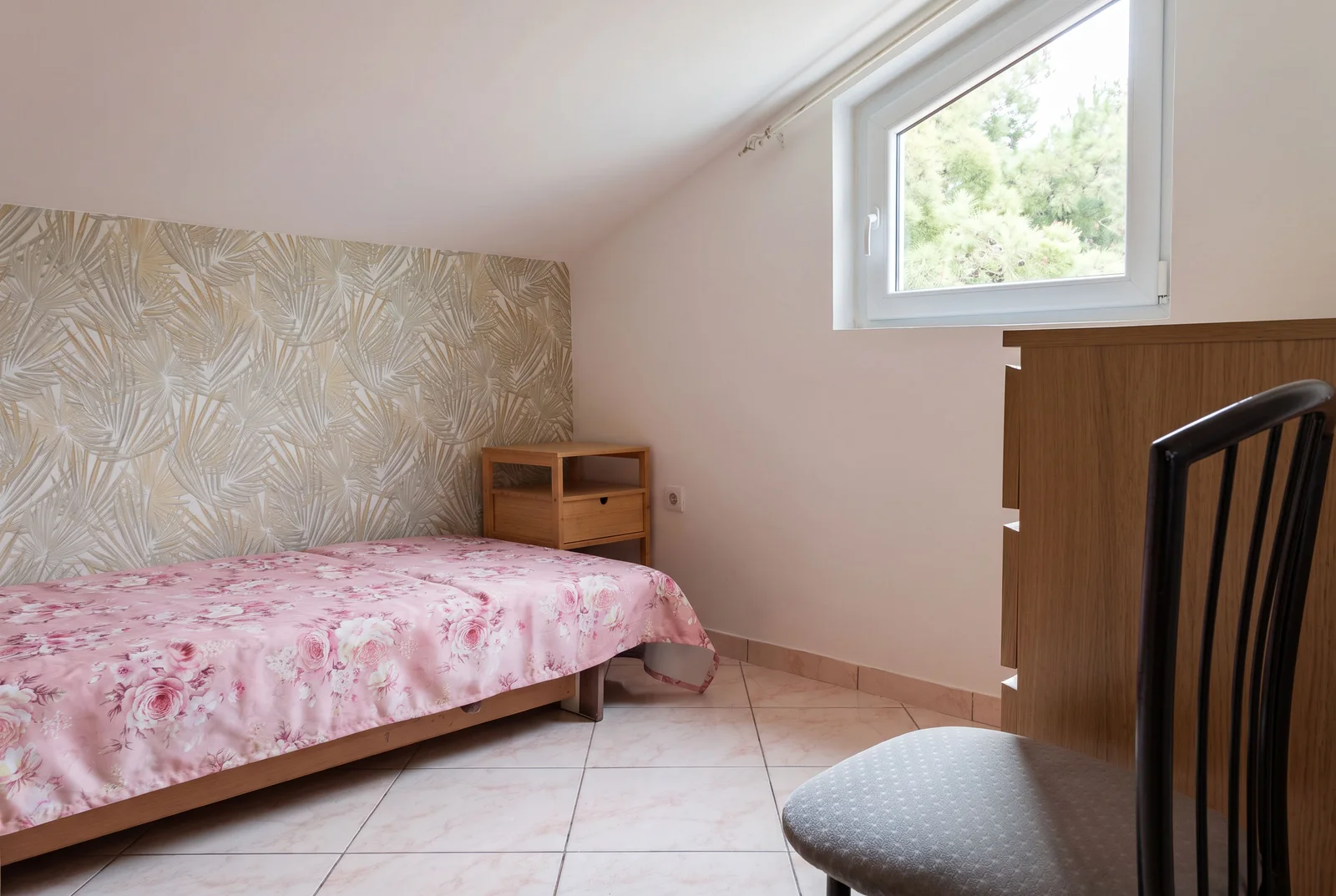 Bedroom with a single bed in floral bedding, wooden nightstand, patterned wallpaper, and a window.
