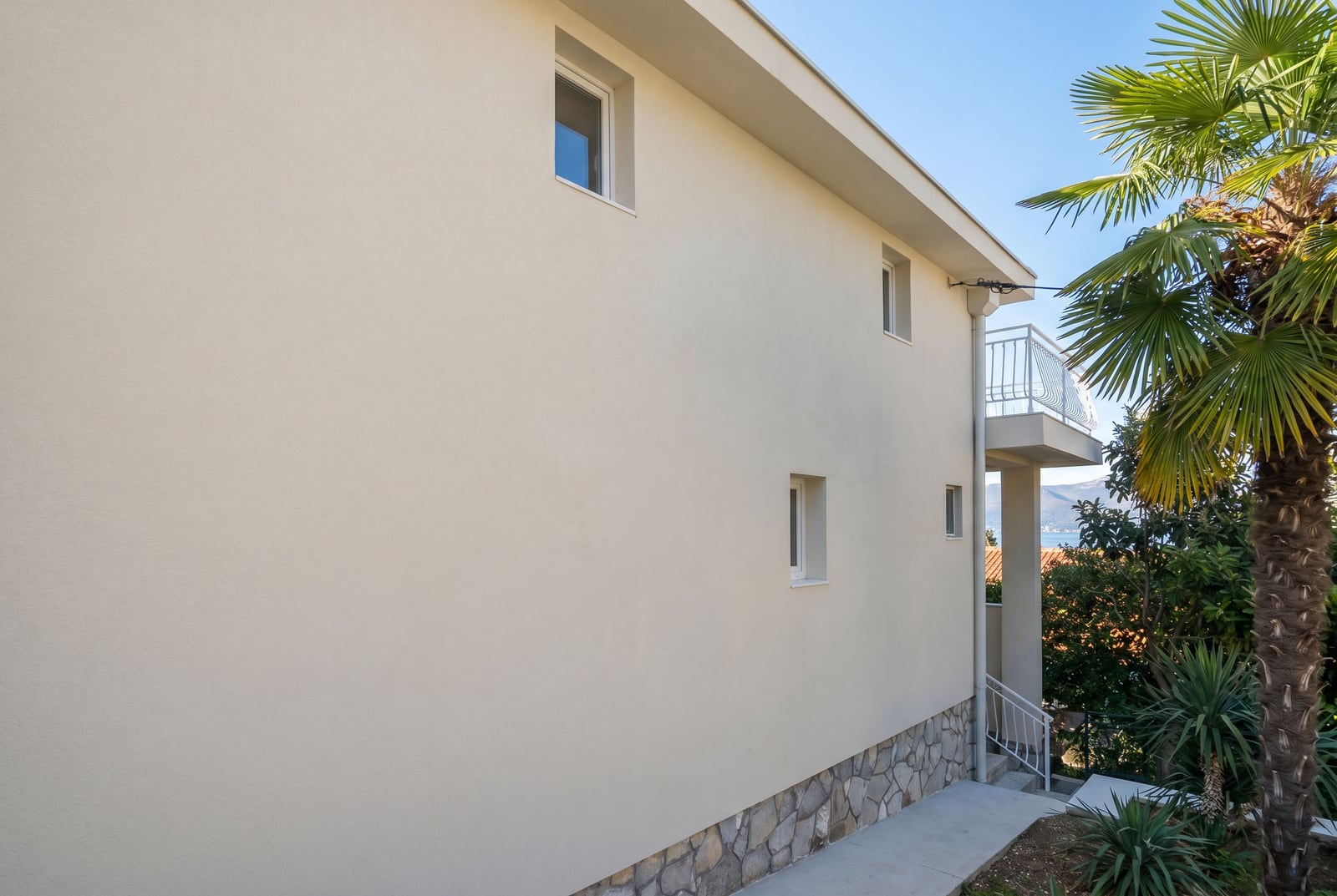 Facade of a two-story house in Krašići, Montenegro, featuring stone base trim, a balcony, and a palm tree on the property.