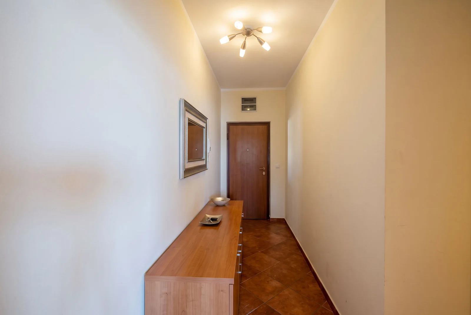 Narrow hallway featuring a wooden console table, framed mirror, and a closed door at the end.
