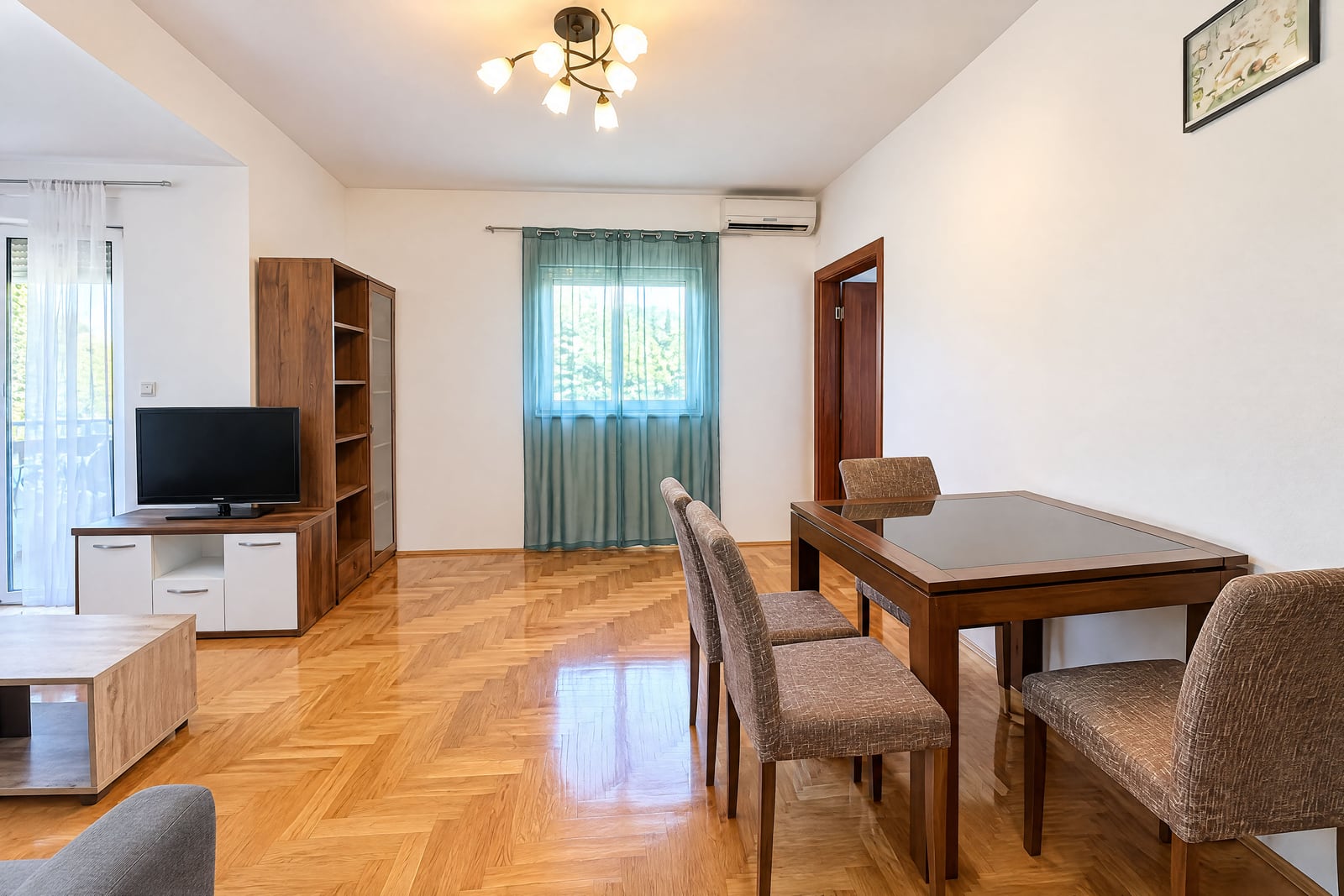 Living room with parquet floor, TV unit, wooden cabinet, and a dining table with upholstered chairs.