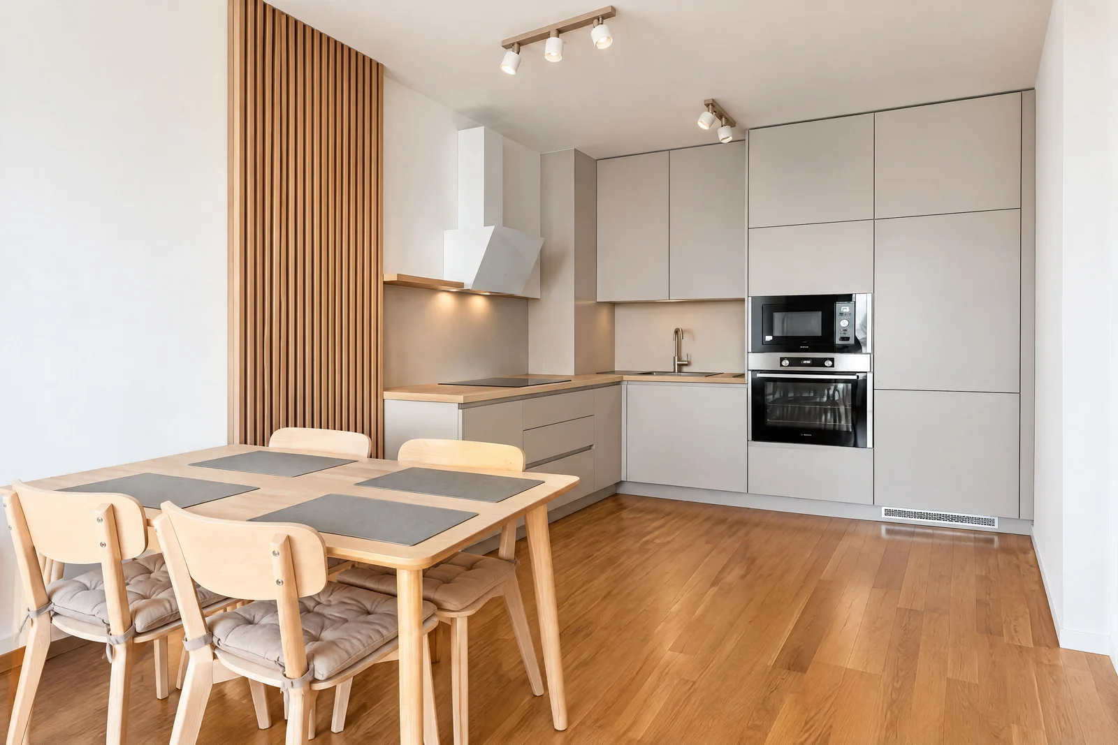 Modern kitchen with light grey cabinets, built-in oven and microwave, wooden dining table, and slatted wood wall accent.