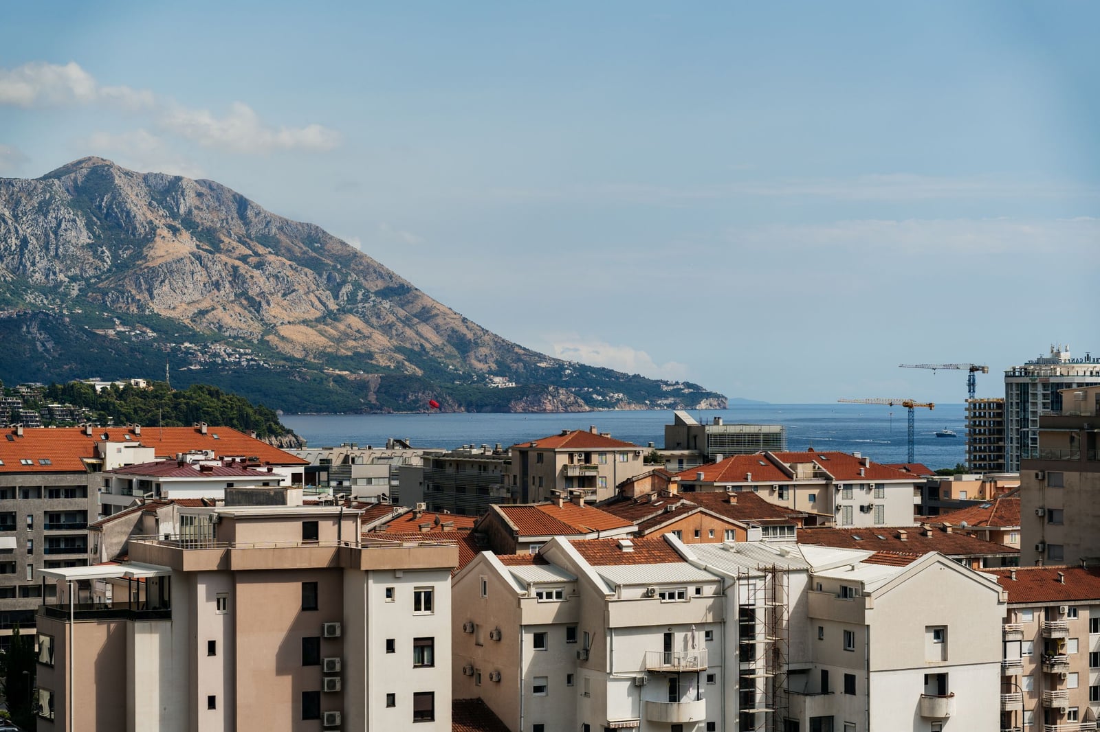 Panoramic view of Budva, Montenegro: residential buildings with tiled roofs, the sea, and a mountain landscape in the background.