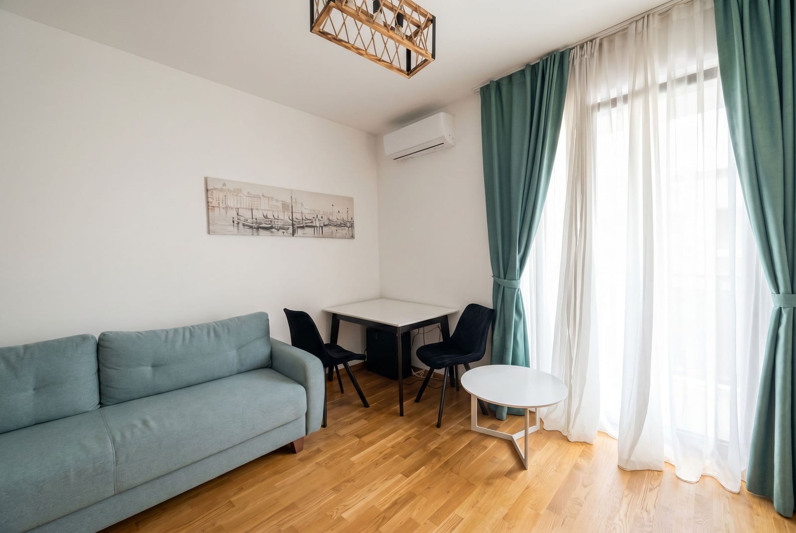 Living room in an apartment in Bečići, Montenegro: sofa, dining area, parquet flooring, and panoramic window with turquoise curtains.