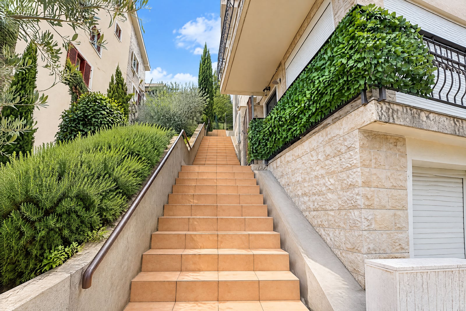 Long outdoor staircase with terracotta tiles leading up between buildings and green bushes under a blue sky.