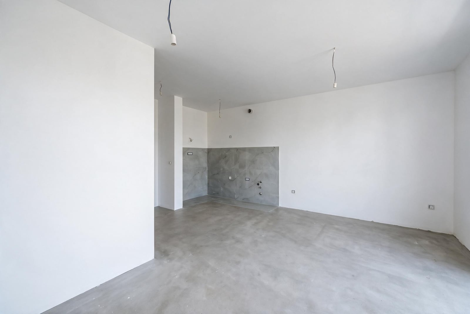 Empty unfinished room with concrete floor and a section of grey wall tiles, featuring hanging electrical wires.