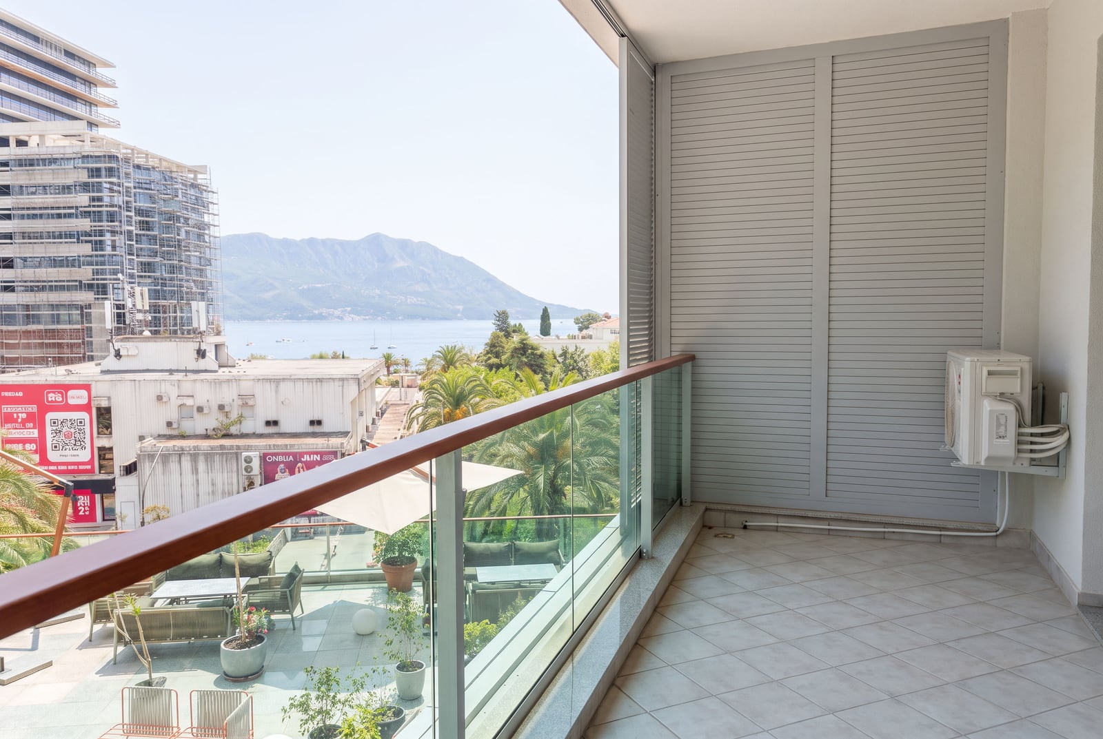 Panoramic balcony with glass railing, sea and mountain view, grey shutters, and wall-mounted AC unit.