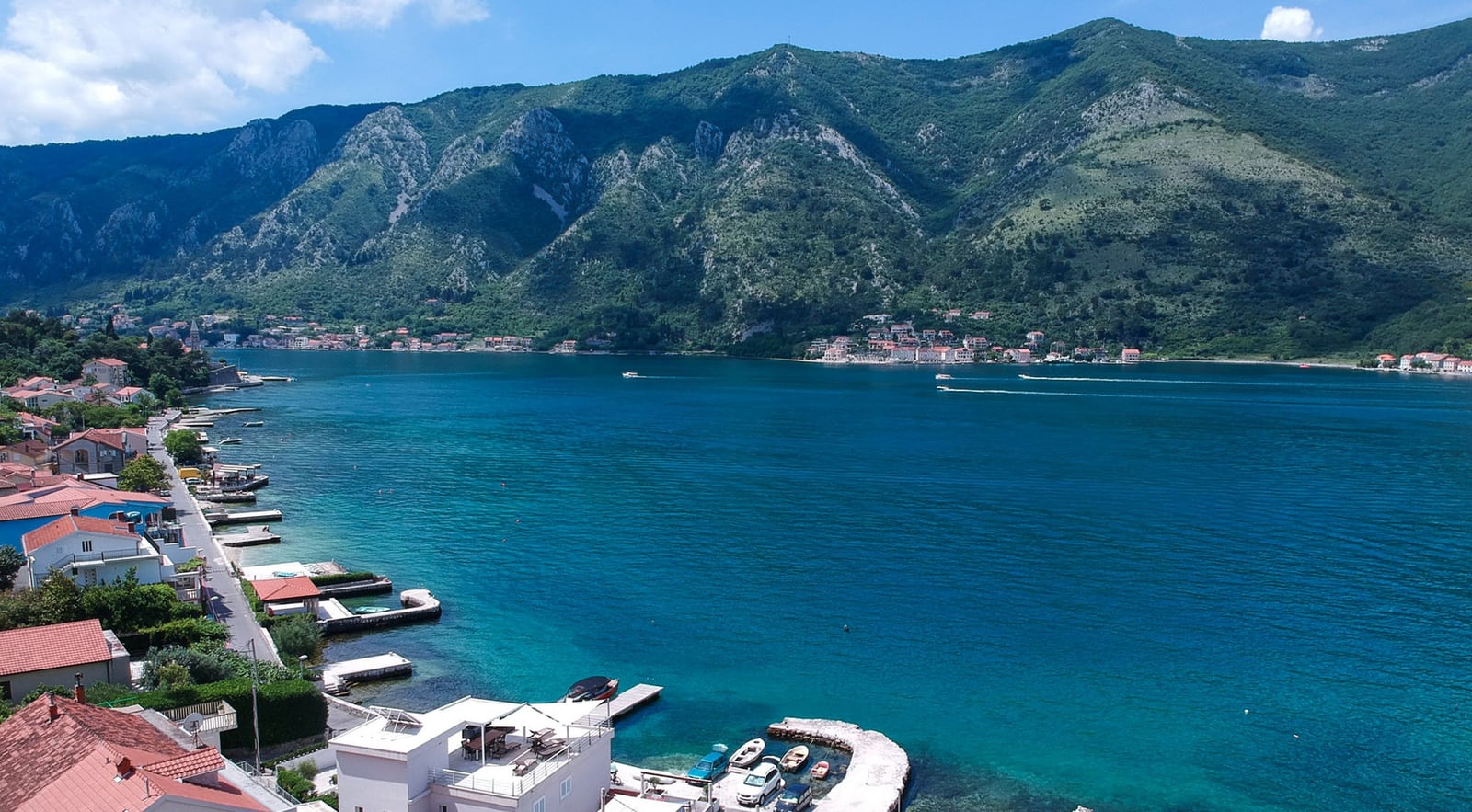 Panoramic view of Kotor Bay and a villa in Dobrota, Montenegro: coastline, piers, and mountains in the background.
