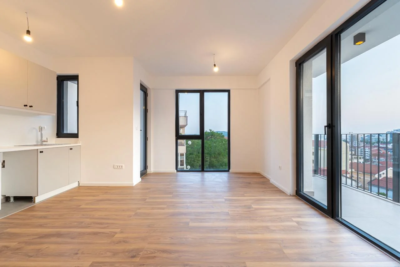 Empty room with laminate flooring, kitchen unit on the left, and large glass doors to a balcony on the right.