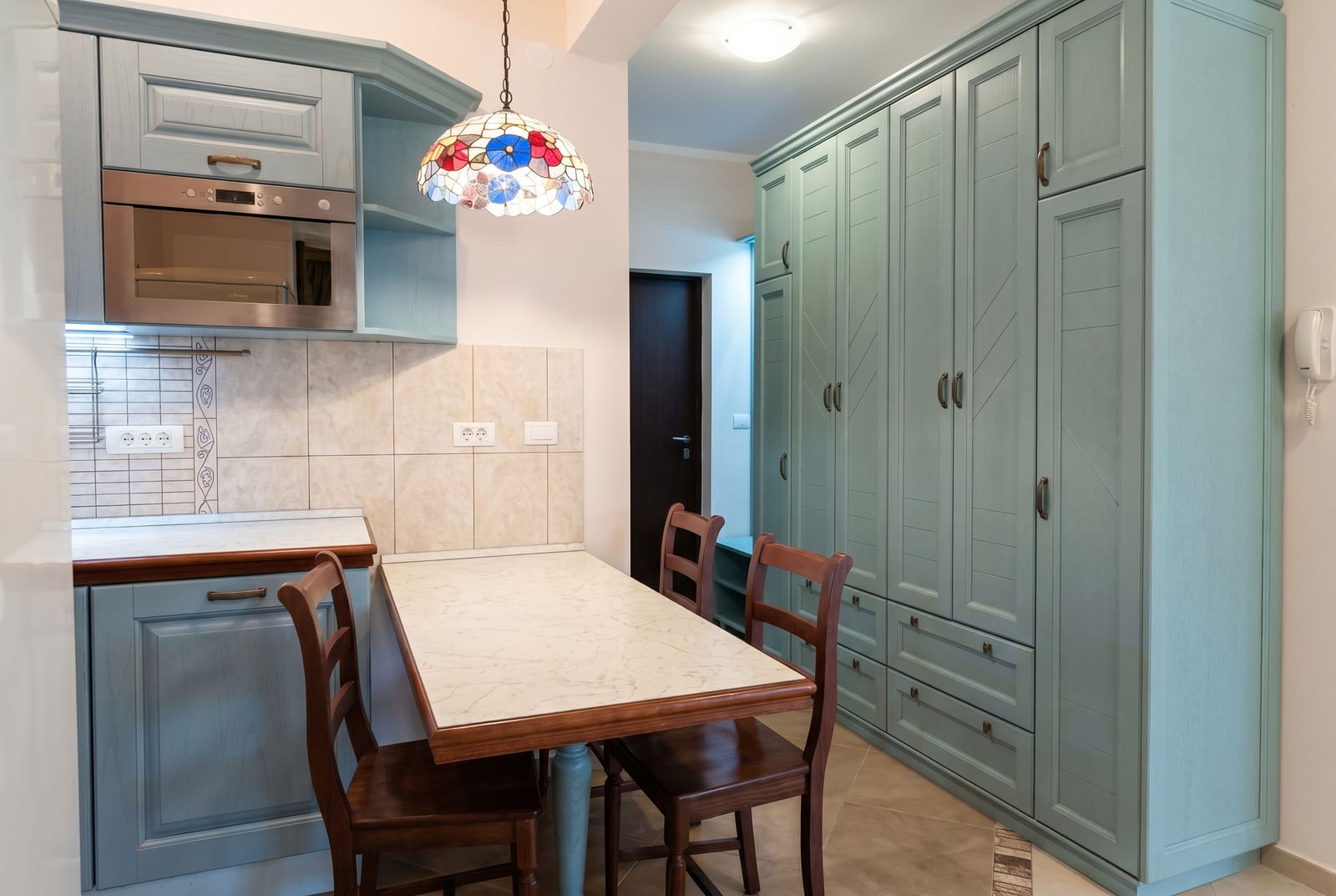 Kitchen in an apartment in Bar, Montenegro: blue cabinetry, dining table, wooden chairs, and a stained-glass lamp.