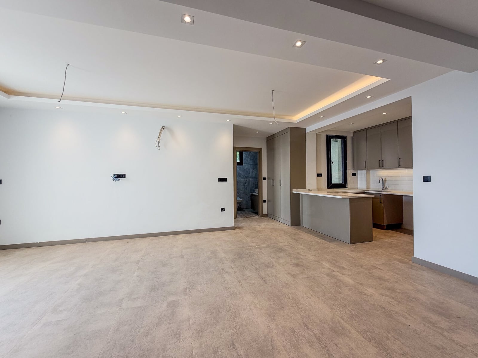 Empty open space with tile flooring and an adjacent kitchen island, exposed wiring hanging from the ceiling.