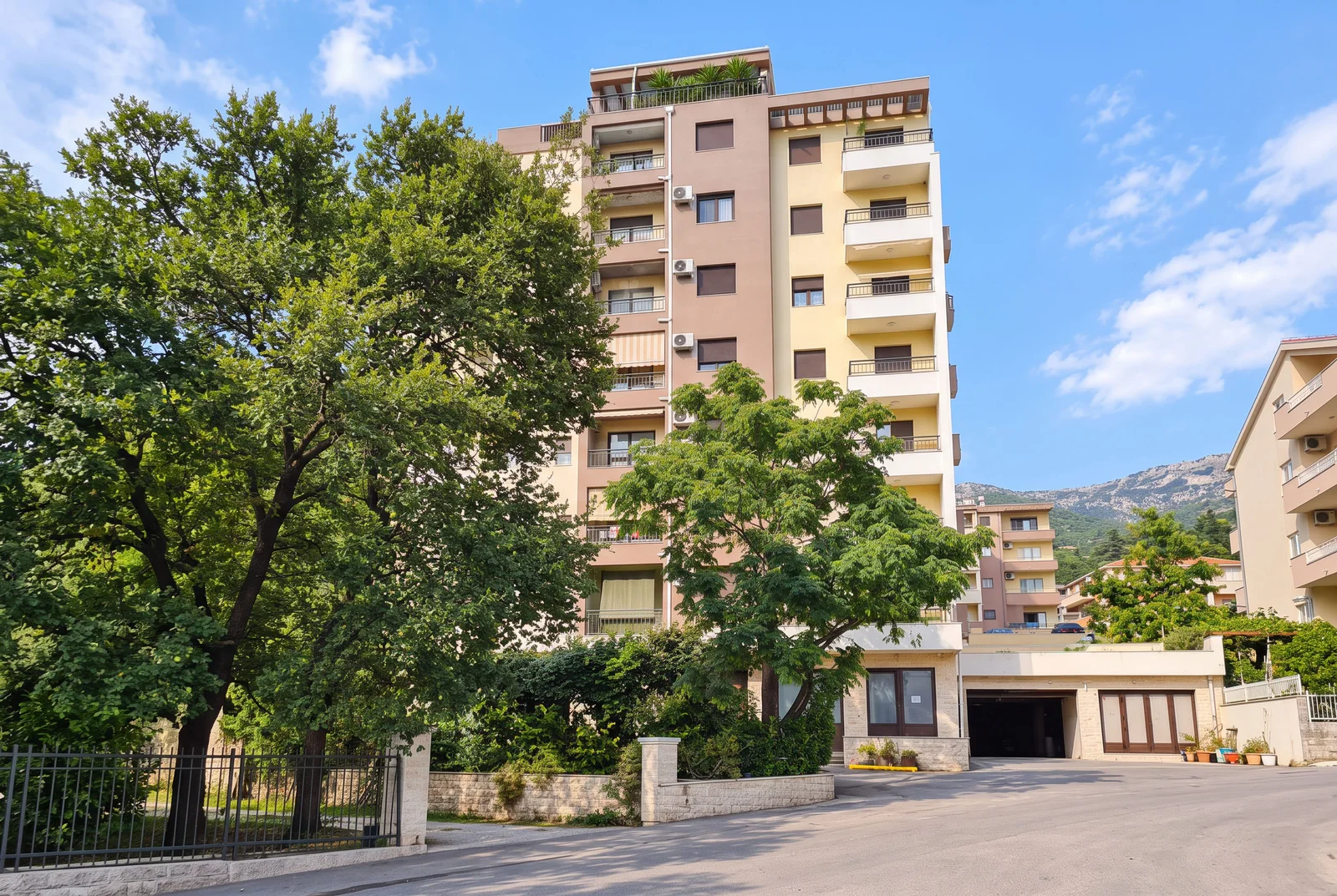 Exterior view of a multi-story residential building with balconies, surrounded by green trees under a blue sky.