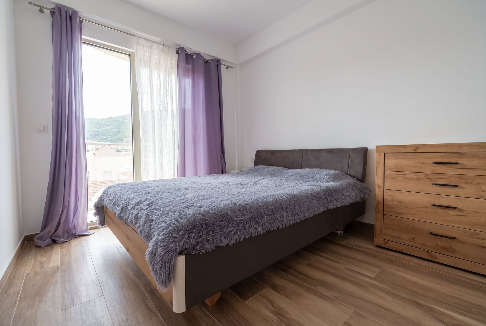 Bedroom featuring a double bed with a grey fluffy cover and a wooden dresser next to a window with purple curtains.