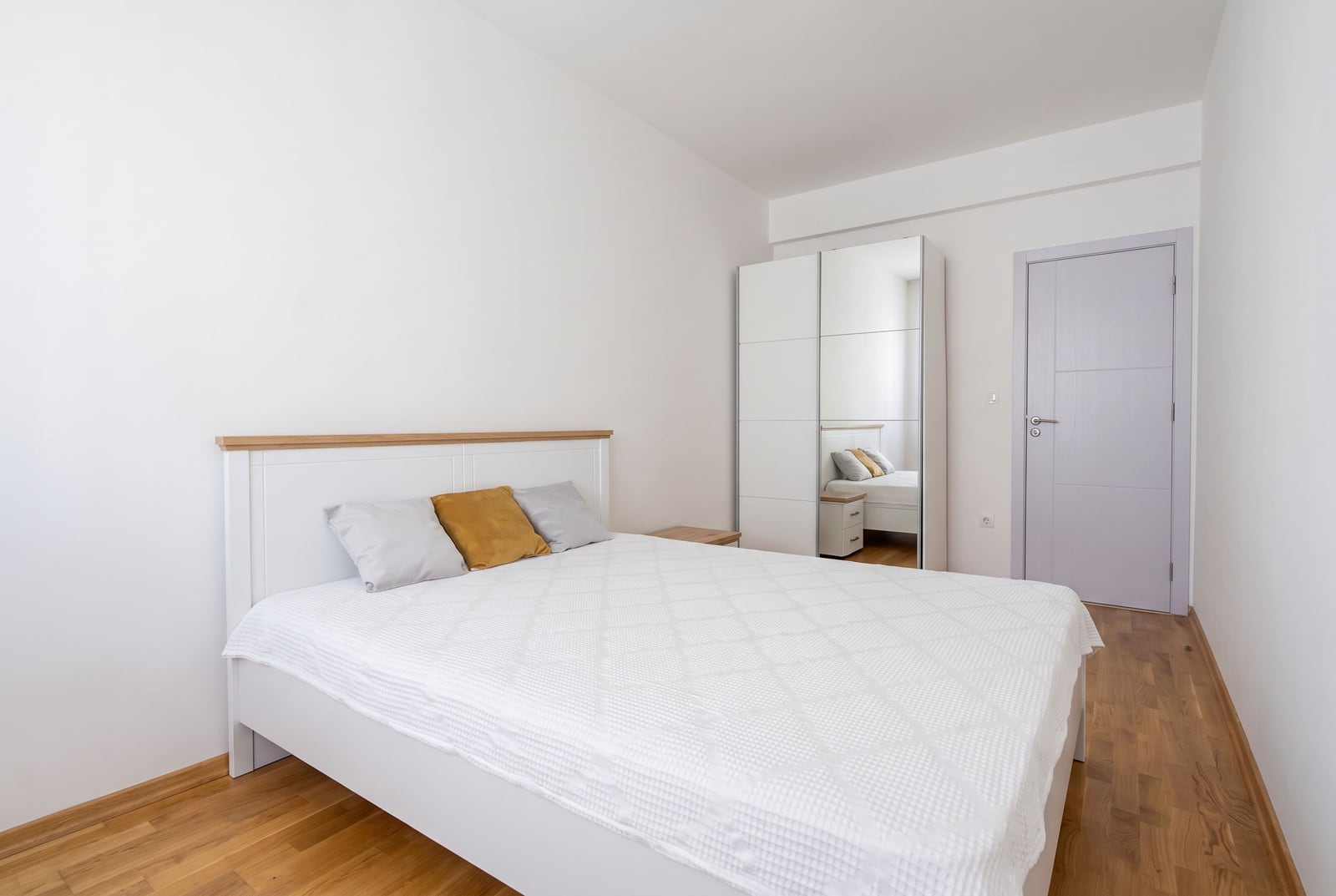 Bright bedroom featuring a white bed with wood headboard, mirrored wardrobe, and wooden parquet flooring.