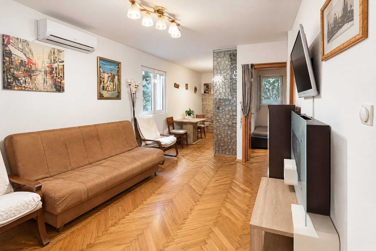 Living room featuring a brown sofa, herringbone wood floor, wall-mounted TV, and a dining area visible in the background.