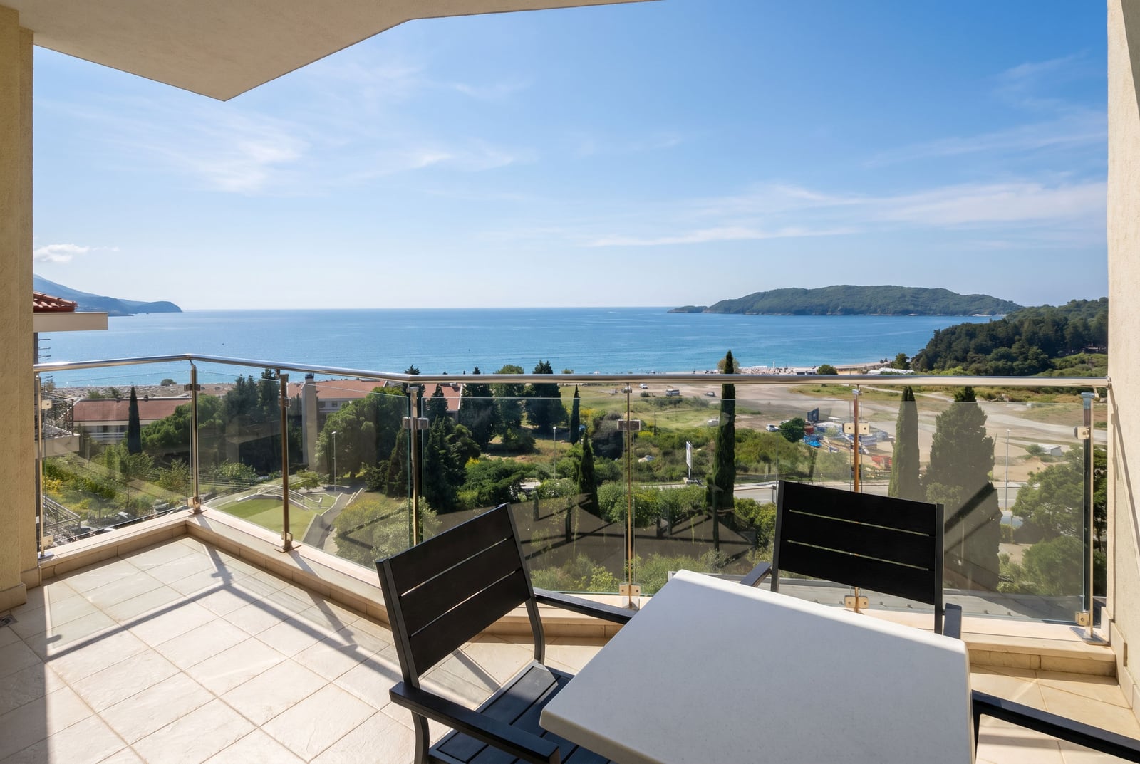 View from the balcony of an apartment in Bečići, Montenegro: a table with chairs, glass railing, and a panorama of the sea and beach.