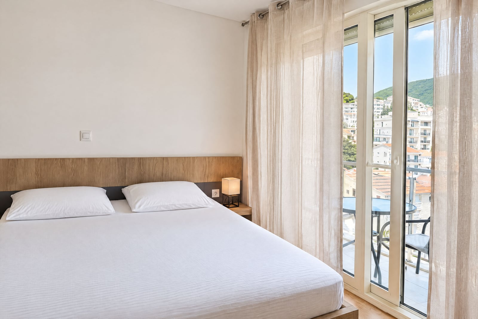 Bright bedroom featuring a double bed with a wooden headboard and glass doors leading to a balcony overlooking the city.