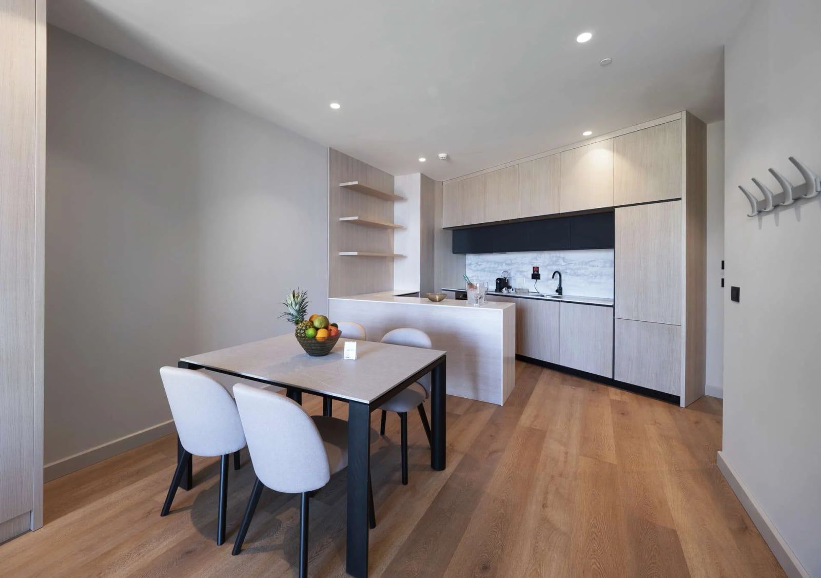 Modern kitchen with light wood cabinets, a white island, dining table with fruit bowl, and wooden flooring.