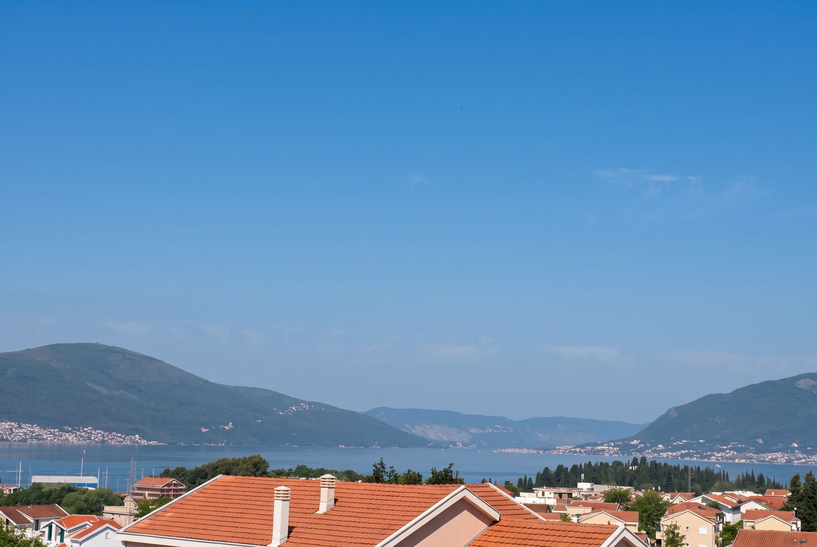 Panoramic view of red tile rooftops, sea, and mountains under a clear blue sky.