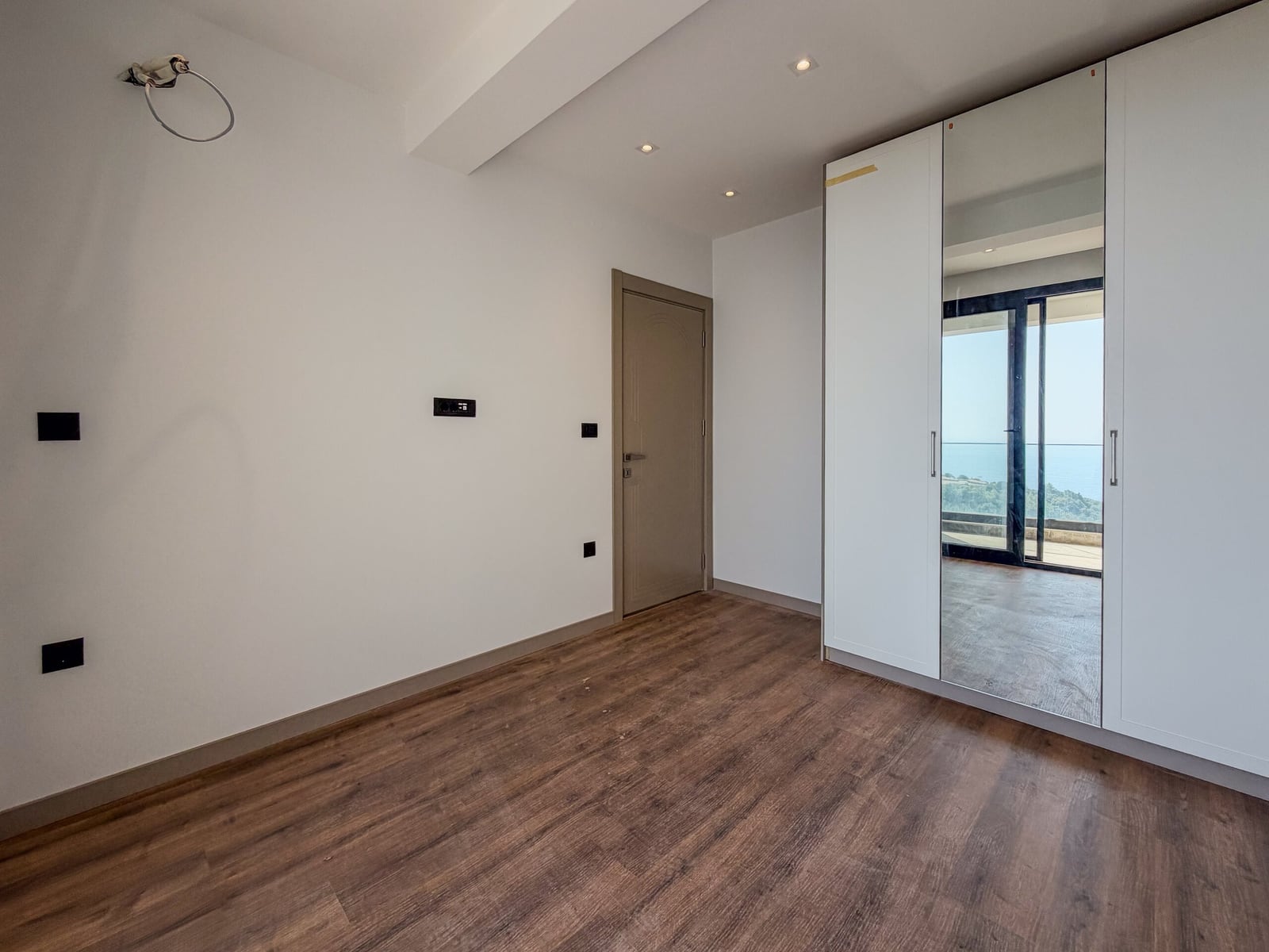Empty room with wood flooring and a white wardrobe with a mirror. A door and wall outlets are visible.