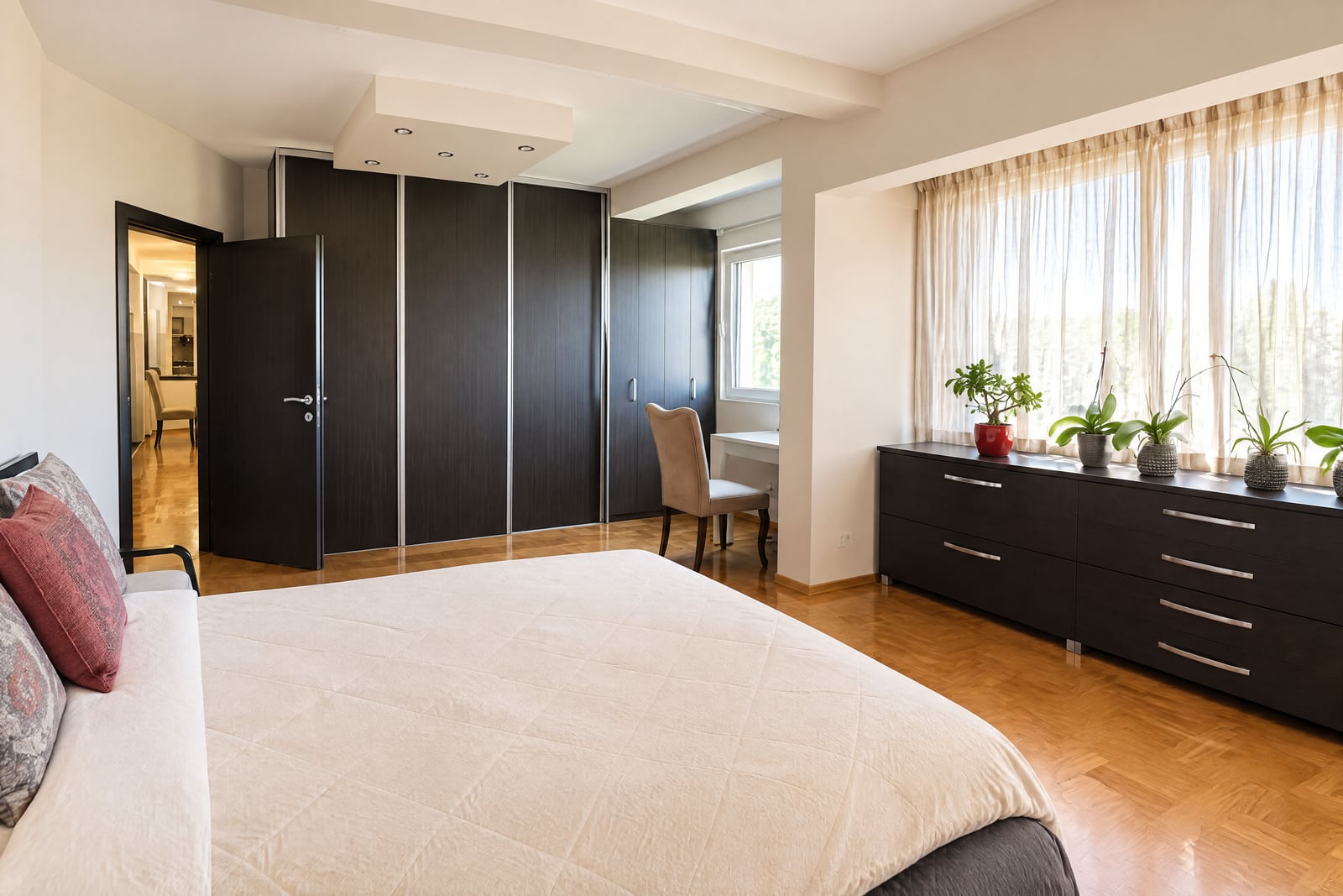Bedroom featuring a large bed, dark sliding wardrobes, a dresser with plants by the window, and parquet flooring.