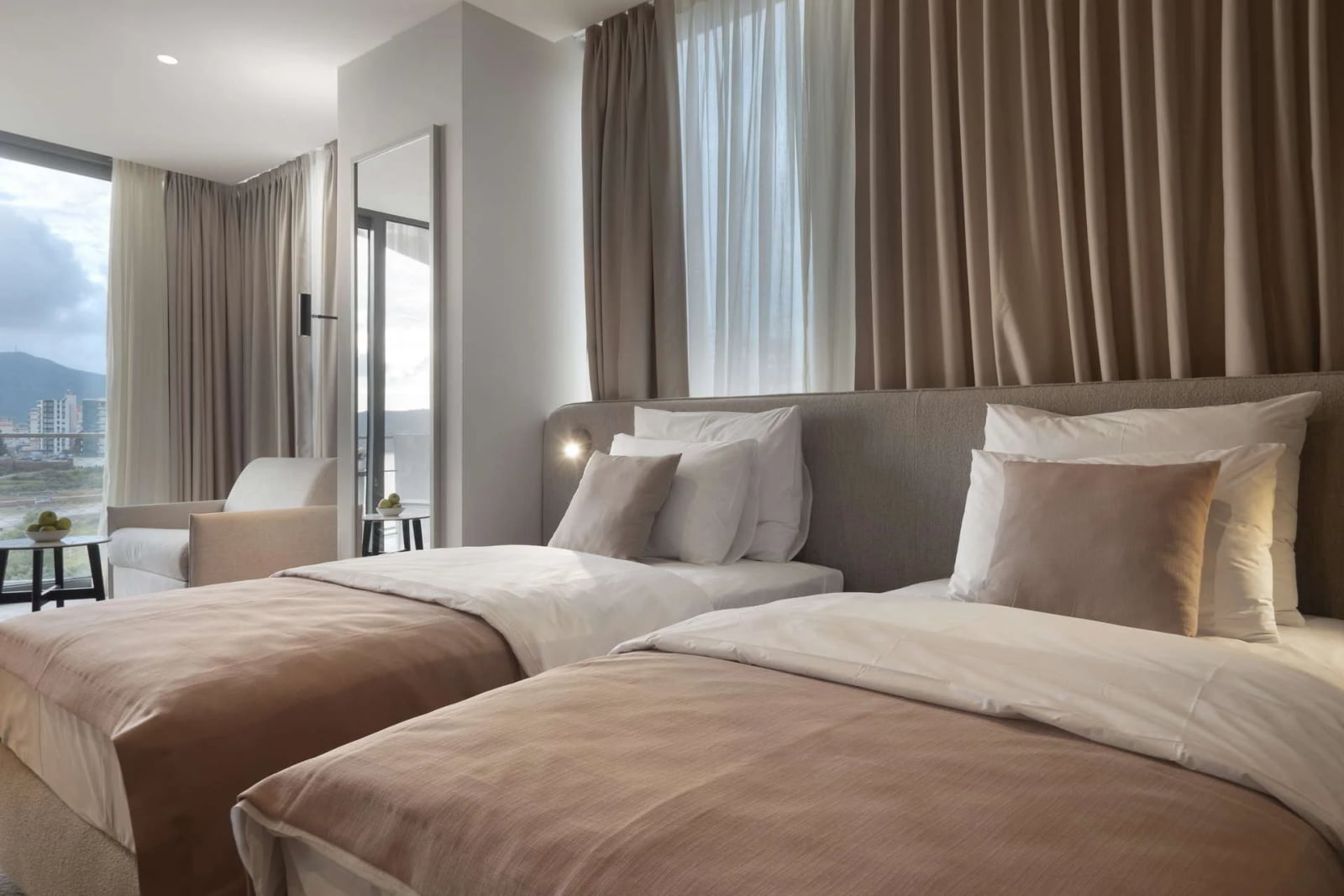 Two single beds with beige bedding, upholstered headboard, floor-length curtains, and an armchair near the window.