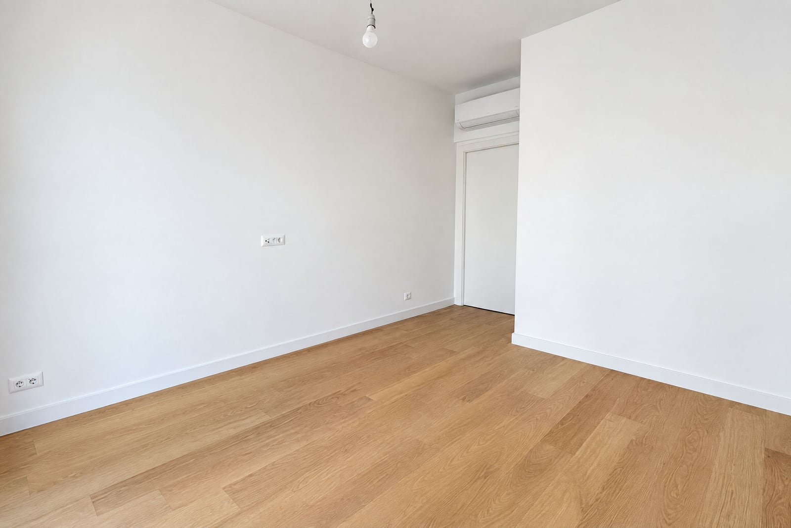 Empty room with white walls, light wood flooring, a hanging bulb, and an air conditioner near the door.