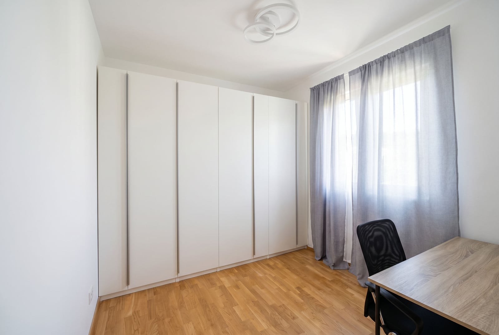 Bedroom in an apartment in Bečići, Montenegro: built-in white wardrobe, desk, parquet flooring, and light-colored curtains on the window.