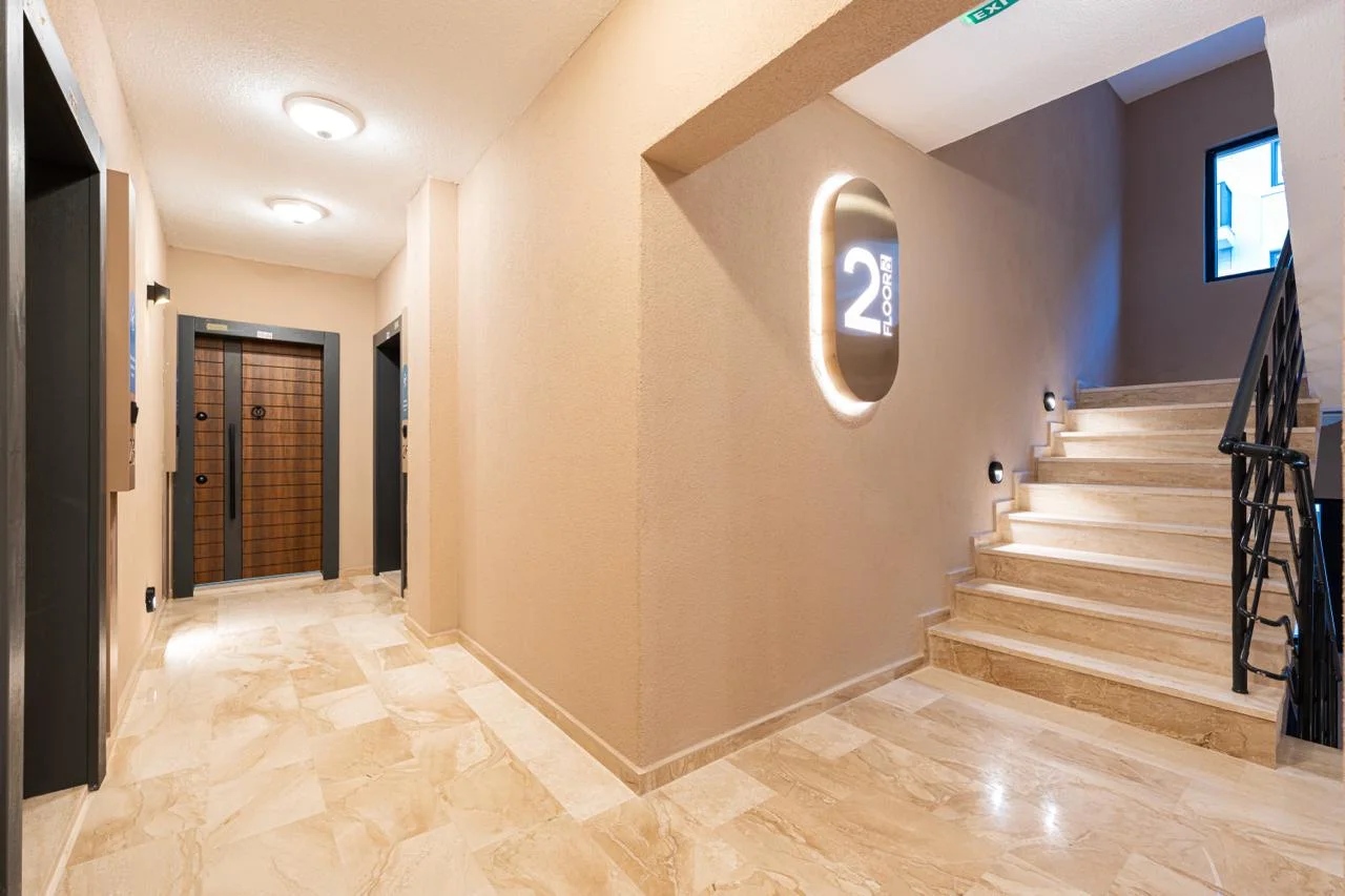 Hallway with staircase, entrance door, and illuminated floor sign on a beige wall.