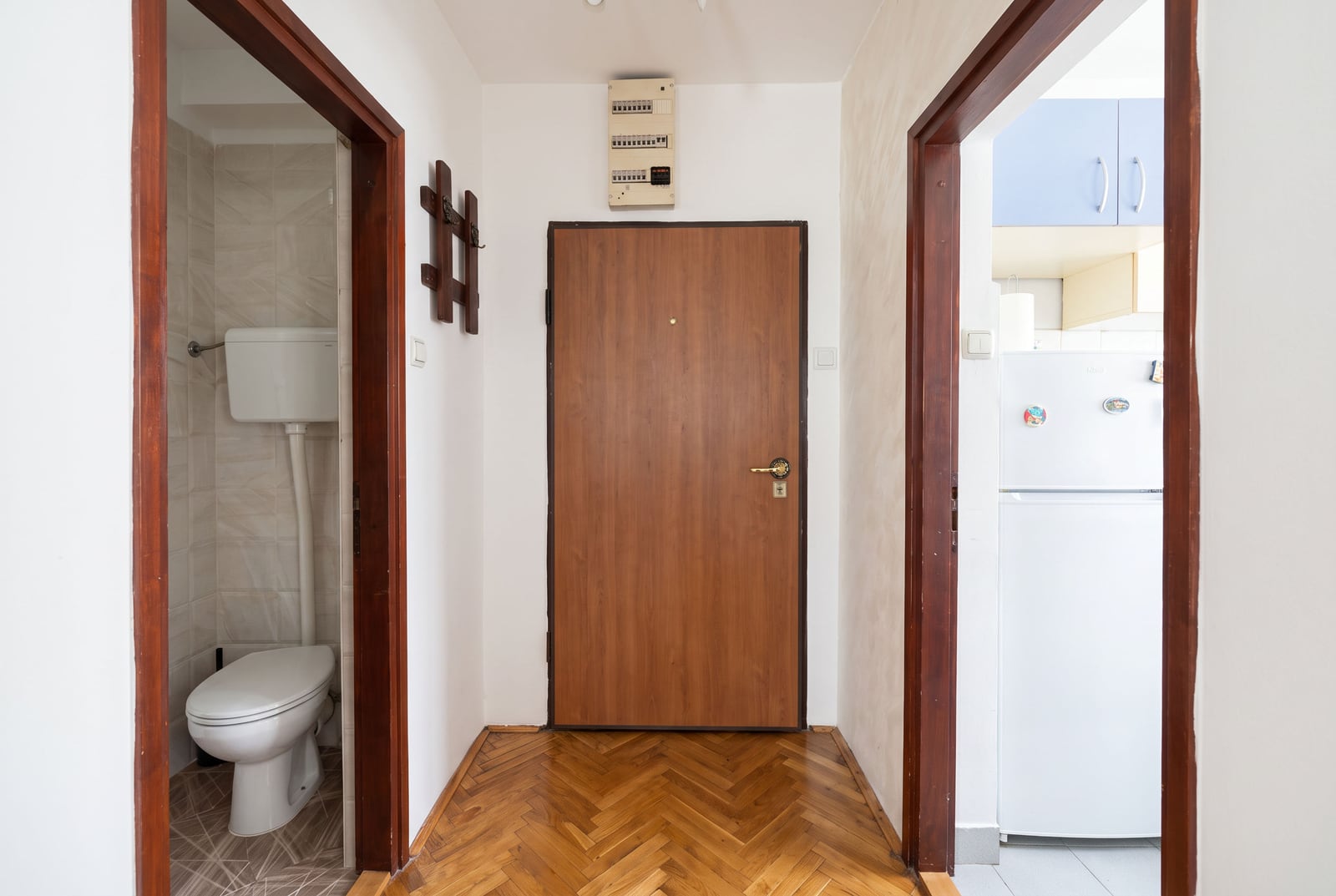 Hallway with herringbone parquet, entrance door, partial views of a toilet and kitchen.